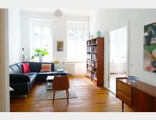 Helles Wohnzimmer mit Holzboden, einer schwarzen Eckcouch mit bunten Kissen, einem kleinen Holztisch und einem Bücherregal. Die weißen Wände sind mit großen Fenstern, einem Gemälde und Zimmerpflanzen dekoriert, nebenan ist ein weiterer Raum sichtbar.