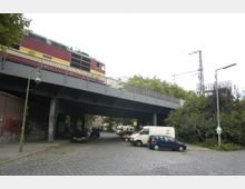 Eine erhöhte Eisenbahnbrücke aus Stahl mit einem vorbeifahrenden Zug darüber. Darunter eine gepflasterte Straße mit geparkten Autos, flankiert von Bäumen und einem Backsteingebäude mit Graffiti.