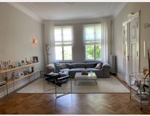 Helles Wohnzimmer mit einem grauen Ecksofa, hellen Vorhängen und großen Fenstern, die Tageslicht hereinlassen. Holzparkettboden, ein schlichter Couchtisch, ein offenes Regal mit dekorativen Elementen und eine weiß gestrichene Tür mit verziertem Rahmen prägen den Raum.