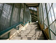 Steintreppe mit grünen Handläufen in einem überdachten Gang mit großen Glasfenstern und Metallrahmen. Der Gang hat ein schräges Dach und bietet Blick auf den Himmel durch die Fenster.