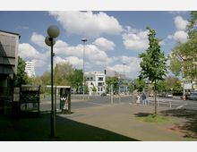 Straßenszene in einer städtischen Umgebung mit einer Kreuzung, Verkehrszeichen und einer Haltestelle mit Wartehäuschen im Vordergrund. Im Hintergrund sind mehrstöckige Gebäude, Bäume und blauer Himmel mit Wolken zu sehen.