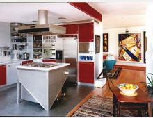 Moderne Küche mit roten und weißen Schränken, einem zentralen Edelstahl-Kochblock mit Dunstabzugshaube und silbernen Elektrogeräten. Im Hintergrund ein angrenzender Wohnbereich mit Holzboden, Teppichen und einem großen bunten Gemälde an der Wand.