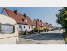 Eine ruhige Wohnstraße mit einer Reihe von Einfamilienhäusern, die rote Ziegeldächer und hell verputzte Fassaden in verschiedenen Farben haben. Im Vordergrund ist ein geparkter Wohnwagen zu sehen, und die Straße führt an Bäumen und parkenden Autos entlang.