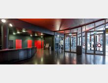 Moderner Eingangsbereich mit großen verglasten Doppeltüren, schwarzen Wänden und Decken sowie roten Türen im hinteren Teil. Links ist ein geschwungener, dunkler Empfangstresen, während der Boden aus reflektierendem dunklem Material besteht.