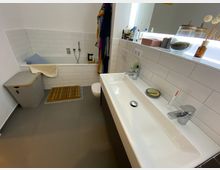 Modernes Badezimmer mit grauen Bodenfliesen, einer Badewanne mit weißer Fliesenverkleidung, einem großen weißen Waschbecken und Aufbewahrungsflächen mit Dekorationen. Daneben steht ein Wäschekorb, und vor der Badewanne liegt eine Holzmatte.