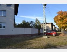 Straßenszene in einem Wohngebiet mit Mehrfamilienhäusern aus Putz und Flachdächern, hellgrauen Mauern und einem vorgelagerten Grünstreifen. Im Vordergrund ein Strommast, daneben ein rotes geparktes Auto und ein Baum mit herbstlich verfärbtem Laub.