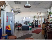 Ein modernes Büro mit hellem, offenen Raum, weißer Decke und Fliesenboden. Im Vordergrund stehen Sitzhocker, ein Kühlschrank mit Red-Bull-Logo, Schreibtische mit Bürostühlen und Computerbildschirmen, sowie Regale mit Dekorationen, darunter Pflanzen und Holzstämme als Gestaltungselemente.