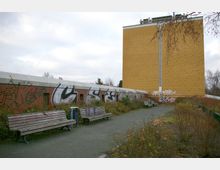 Ein urbaner Außenbereich mit mehreren Holzbänken entlang eines gepflasterten Weges, umgeben von Sträuchern und Blumen. Im Hintergrund ein gelbes, hohes Gebäude mit einer glatten Fassade und ein niedrigeres Backsteingebäude mit Graffiti.