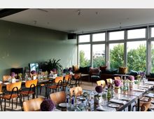 Ein modern eingerichteter Veranstaltungsraum mit langen Holztischen und hellbraunen Stühlen, dekoriert mit lila Blumen und gedecktem Geschirr. Große Fenster an einer Seite des Raumes bieten Ausblick auf grüne Bäume; daneben befindet sich eine Sitzbank mit bunten Kissen.