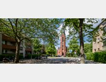 Eine Backsteinkirche mit hohem Turm und Spitzdach steht zentral in einer von Bäumen gesäumten Straße, flankiert von Wohngebäuden. Im Vordergrund befinden sich Gehwege, parkende Fahrräder und Bänke, während Personen den Kircheneingang passieren.