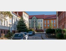 Straßenszene mit gepflastertem Weg, flankiert von mehrstöckigen Wohngebäuden mit roten Ziegeldächern in verschiedenen Farben (weiß, grün und orange). Links sind geparkte Autos und einige Bäume, während rechts ein rotes Backsteingebäude sowie Büsche und kleine Grünflächen zu sehen sind.