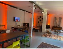 Ein moderner Veranstaltungsraum mit offenem Grundriss, sichtbaren Deckenbalken und weißen Säulen. Der Raum ist mit verschiedenen Tischen, Stühlen in bunten Farben, Teppichen und Wanddekorationen eingerichtet; eine dezente orangene Beleuchtung schafft eine warme Atmosphäre.