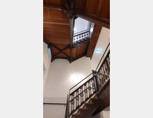 Holztreppenhaus mit verziertem Geländer und sichtbaren tragenden Holzbalken unter der Decke. Die helle Wand und eine stilvolle Wandlampe ergänzen die Zwischengeschosse, während ein Notausgangsschild in der oberen Ecke sichtbar ist.