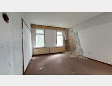 Ein leerer, verlassener Raum mit abgenutztem Holzboden und teils beschädigten Wänden; eine Wand zeigt freiliegendes Mauerwerk und abblätternden Putz. Zwei Fenster mit Heizkörpern darunter lassen Tageslicht herein, und der Raum wirkt unrenoviert.