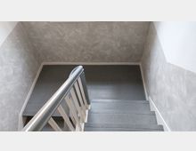 Treppenhaus mit grauen Stufen, einem silberfarbenen Handlauf und Wänden in grauer Putzoptik. Die Treppe führt zu einem kleinen Zwischenpodest.