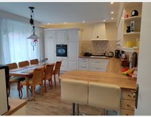 Geräumige Wohnküche mit hellen Holzmöbeln und weißen Küchenschränken. Im Vordergrund eine Küchentheke mit Barhockern, im Hintergrund ein Essbereich mit einem großen Holztisch und Stühlen sowie eine Kochzeile mit integriertem Backofen und Dunstabzugshaube.