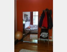 Zimmer mit orangefarbener Wand, einem Kleiderschrank mit Spiegeltüren sowie einem Holzfußboden. Im Vordergrund steht ein Stuhl mit einer Sporttasche, daneben hängt eine Garderobe mit Jacken. Im Spiegel ist ein Bett, ein Fenster mit Vorhängen und ein Heizkörper zu sehen.