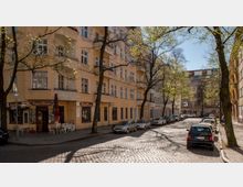 Straßenszene in einem Wohnviertel mit Mehrfamilienhäusern aus gelbem Putz und anliegenden Balkonen; gepflasterte Straße mit parkenden Autos und Bäumen, deren junge Blätter im Sonnenlicht leuchten. Im Erdgeschoss befindet sich ein Café mit Außenmöbeln.