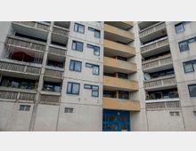 Mehrstöckiges Wohngebäude aus Beton mit beige-grauer Fassade, blauen Fensterrrahmen und vergitterten Balkonen. Im Erdgeschoss befindet sich ein blauer Eingang, einige Balkone sind mit Pflanzen und Dekoration geschmückt.