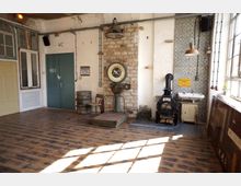 Raum mit industriellem Vintage-Charme, ausgestattet mit Holzboden, unverputzten Ziegelwänden und einer Digitalwaage vor einer gemusterten Fliesenwand. Im Hintergrund befinden sich eine grüne Metalltür mit "WC"-Beschriftung, ein kleiner Kaminofen und ein Waschbecken.