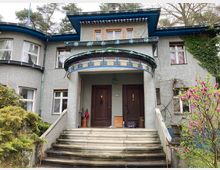 Historisches Wohnhaus mit grauer Fassade, geschwungenen Bauformen und farbigen Akzenten am Vordach. Der Eingangsbereich wird über eine breite Treppe erreicht, umgeben von Bäumen, Sträuchern und einer blühenden Magnolie.