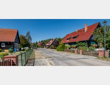 Straßenszene in einem Wohngebiet mit Einfamilienhäusern, die rote Ziegeldächer und dunkle Fassaden haben. Die Straße ist asphaltiert, gesäumt von Gärten mit Zäunen, und im Hintergrund sind Bäume sowie ein klarer blauer Himmel sichtbar.