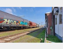 Ein Güterzug mit Graffiti auf den Waggons fährt auf einem Gleis neben einem roten Ziegelgebäude mit einzelnen Graffiti. Im Hintergrund sind weitere Schienen und Bahninfrastruktur zu sehen, darunter Oberleitungen und Lampenmasten.