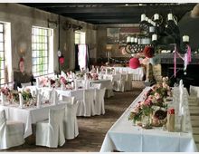 Rustikaler Veranstaltungsraum mit großen Fenstern und dunklen Holzbalkendecken, eingedeckt mit weiß gedeckten Tischen für eine Feier. Der Raum ist mit bunten Blumenarrangements, Kerzen und hängenden Papierdekorationen geschmückt.