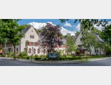 Zweistöckiges Gebäude im historischen Stil mit heller Fassade und Satteldach, umgeben von Bäumen und gepflegten Gärten. Vor dem Gebäude steht eine Informationswand, und im Hintergrund sind weitere Grünflächen sowie ein kleiner Turm zu sehen.