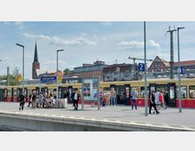 Ein S-Bahnhof in Berlin mit einem gelb-roten S-Bahn-Zug an Gleis 2. Menschen stehen und gehen auf dem Bahnsteig, im Hintergrund sind eine Backsteinfassade mit Graffiti, ein Kirchturm und weitere Gebäude zu sehen.