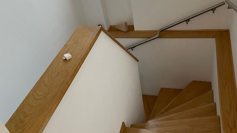 Innenansicht eines hellen Treppenhauses mit Holzstufen und Holzgeländern, das nach unten führt. Weiße Wände, ein Metallhandlauf an der rechten Seite und ein schmales Fenster sorgen für Tageslicht.