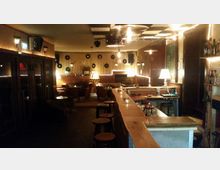 Gemütliche Bar mit Thekenbereich aus Holz und mehreren Hockern, umgeben von Lounge-Sesseln und Tischen. Die Wände sind mit Schallplatten dekoriert, Lampen sorgen für eine warme, atmosphärische Beleuchtung.