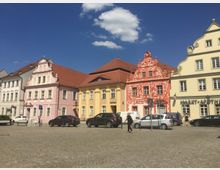 Reihe von historischen Gebäuden mit bunten Fassaden in Pastelltönen (rosa, gelb und rot) mit dekorativen Giebeln und roten Dächern, im Vordergrund ein gepflasterter Platz mit geparkten Autos und Personen. Der Himmel ist klar und blau mit wenigen Wolken.