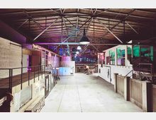 Weitläufiger Innenraum einer umgebauten Industriehalle mit hohen Holzdecken und sichtbarem Stahlträgerwerk. Die Seitenwände sind mit anliegenden Räumen und bunt beleuchteten Glasfenstern versehen; in der Mitte befinden sich ein großer, offener Bereich und künstlerische Installationen.