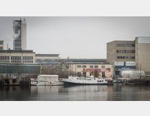 Industriegebiet am Wasser mit mehreren gewerblich genutzten Gebäuden und einem kleinen Hafenbereich. Im Vordergrund liegen Boote an einem ruhigen Gewässer, während im Hintergrund Lagerhallen, Firmenschilder und ein Schornstein sichtbar sind.