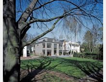 Moderne Wohnhausanlage mit einer Fassade aus Backstein und großen Fenstern, umgeben von einem gepflegten Garten mit Rasen und Bäumen; im Vordergrund ein großer Laubbaum, dessen Äste Schatten auf den Boden werfen. Der Himmel ist klar und blau.