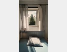 Ein kleines Schlafzimmer mit einem Einzelbett, das direkt unter einem Fenster steht. Das Fenster hat halb geöffnete, lichtdurchlässige Vorhänge und bietet einen Blick auf einen Baum und benachbarte Gebäude.