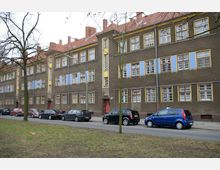 Straßenansicht eines mehrstöckigen Wohngebäudes mit brauner Fassade, gelben und blauen Dekorationselementen sowie roten Dachziegeln. Vor dem Gebäude stehen Autos entlang einer Straße, und im Vordergrund sind Bäume und eine Grünfläche zu sehen.