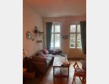 Gemütliches Wohnzimmer mit grauem Ecksofa, niedrigem Couchtisch und Holzstuhl auf Parkettboden. Große, hohe Fenster und eine Balkontür mit Vorhängen lassen Tageslicht herein; an der Wand hängen Regale mit Pflanzen und Dekoration.