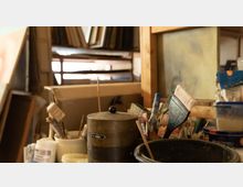 Ein Atelier mit verschiedenen Malutensilien, darunter Pinsel, Behälter und Farben. Im Hintergrund sind Regale mit Bilderrahmen zu sehen.