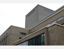 Außenansicht eines mehrstöckigen Gebäudes aus Beton mit rauer Fassade, rechteckigen Fenstern und einer klaren, geometrischen Architektur. Die verschiedenen Ebenen des Gebäudes sind durch horizontale Linien und abgetreppte Strukturen gekennzeichnet.