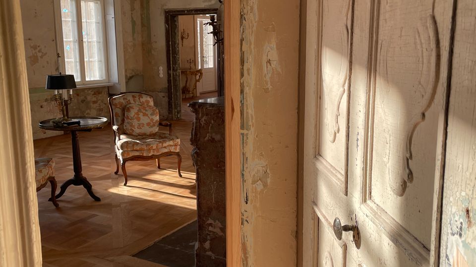 Blick durch eine geöffnete, verzierte Holztür in einen alten Salon mit hohen Fenstern, Parkettboden und einem Kronleuchter. Im Raum stehen ein kleiner runder Tisch, ein gepolsterter Sessel und ein Spiegel; die Wände wirken abgenutzt und teilweise abgeblättert.