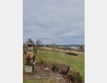 Ein Garten mit einer gemauerten Außengrillecke im Vordergrund, umgeben von kahlen Sträuchern und Rasenflächen. Im Hintergrund ist ein ruhiger See mit umliegender Landschaft und bewölktem Himmel zu sehen.