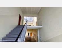 Innenraum eines Treppenhauses mit hellbeigen Wänden, grauen Treppenstufen und einem Metallgeländer. Im oberen Bereich befindet sich ein Lichtschacht aus Glas, darunter führt eine Etage mit Türen und Geländer entlang.