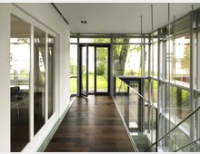 Moderner Flur mit großen Glasfenstern und -türen, die den Blick auf einen gepflegten Garten mit Bäumen und Rasen bieten; dunkler Holzfußboden und eine Glasbrüstung ergänzen das helle, minimalistische Design.