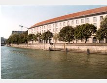 Ein großes, mehrstöckiges Gebäude mit hellem Putz und rotem Ziegeldach steht am Ufer eines Flusses, gesäumt von einer Reihe von Bäumen. Im Vordergrund befindet sich das Wasser des Flusses, und am Ufer sind Poller und eine kleine Anlegestelle sichtbar.