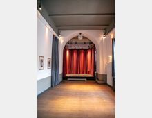 Ein kleiner Veranstaltungsraum mit Holzboden, einer Bühne mit roten Vorhängen und einem Rundbogen darüber. An den Wänden hängen Bilder, und der Raum ist mit dezenten Deckenstrahlern beleuchtet.