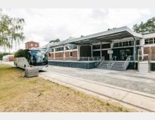 Backstein-Gebäude mit modernem Anbau und überdachtem Eingangsbereich mit Treppen und Rampe, daneben ein geparkter Reisebus auf einer gepflasterten Fläche. Im Vordergrund eine Grasfläche und im Hintergrund Bäume sowie ein weiteres Gebäude.