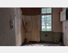 Verlassener Raum mit abgenutzten gelblichen Wänden, teils abblätternder Tapete, und einem großen Fenster mit Blick auf grüne Bäume. Der Boden besteht aus schwarz-weiß karierten Fliesen, und an der rechten Wand befindet sich ein alter Heizkörper.