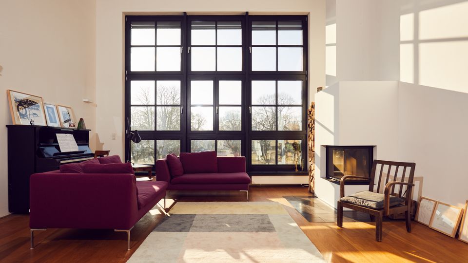 A modern living room with large floor-to-ceiling windows divided into black-framed panes, allowing natural light to fill the space. The room features maroon sofas, a piano with framed artwork on top, a geometric area rug on a wooden floor, and a fireplace with stacked firewood nearby.